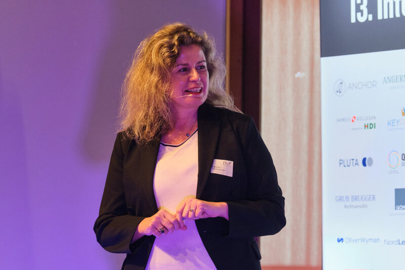 Vortragende Brigitte Kurz steht auf einer Bühne und hält einen Vortrag, im Hintergrund sieht man Sonsorenlogos und den Schriftzug "13. Internationales Symposium Restrukturierung"