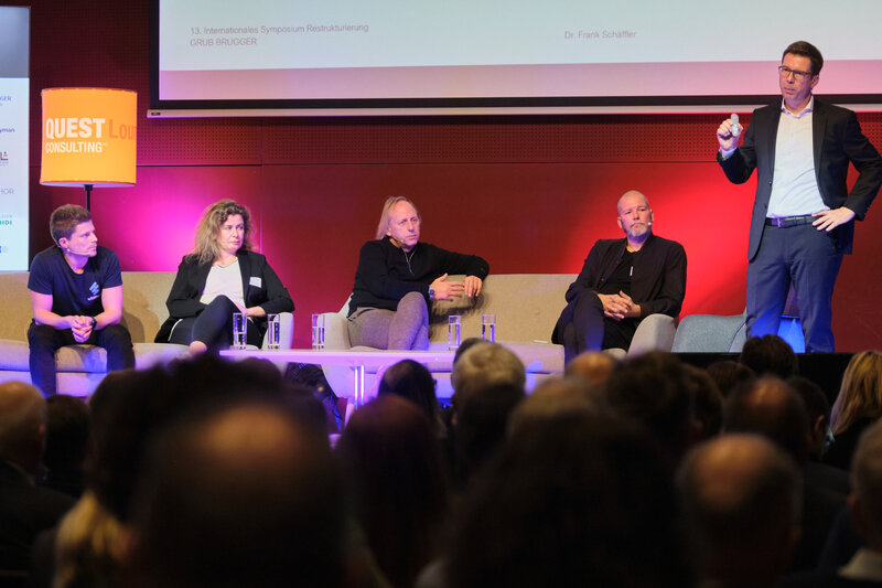 Diskussionsrunde auf einer Bühne, Dr. Frank Schaeffler hält einen Vortrag, auf der Bühne verfolgen eine Frau und drei Männer den Vortrag