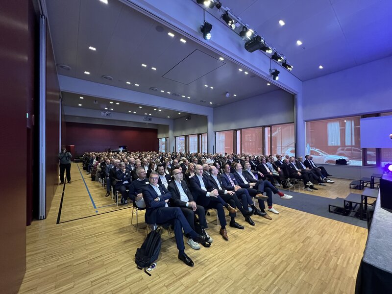 Großer bestuhlter Veranstaltungssaal mit vielen Teilnehmer:innen, am Bildrand ist der Rand einer Bühne zu sehen, das Publikum verfolgt aufmerksam die Veranstaltung
