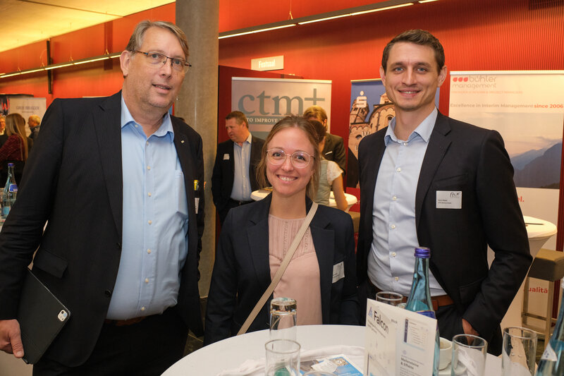drei Besuchende des 13. Internationalen Symposium Restrukturierung, im Hintergrund sind weitere Personen zu sehen, außerdem Aufsteller mit Firmennamen und Logos