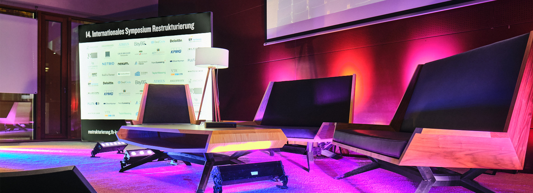 Bühne der 14. Jahreskonferenz Restrukturierung an der HOK, eine moderne Sitzgruppe und mit Tisch und Stehlampe, dahinter eine Aufsteller mit dem Schriftzug 14. Internationales Symposium Restrukturierung und vielen Logos. Die Bühne wird von lila- rosa-farbigen Scheinwerfern angestrahlt