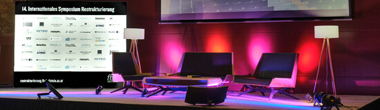 Bühne der 14. Jahreskonferenz Restrukturierung an der HOK, eine moderne Sitzgruppe und mit Tisch und Stehlampe, dahinter eine Aufsteller mit dem Schriftzug 14. Internationales Symposium Restrukturierung und vielen Logos. Die Bühne wird von lila- rosa-farbigen Scheinwerfern angestrahlt