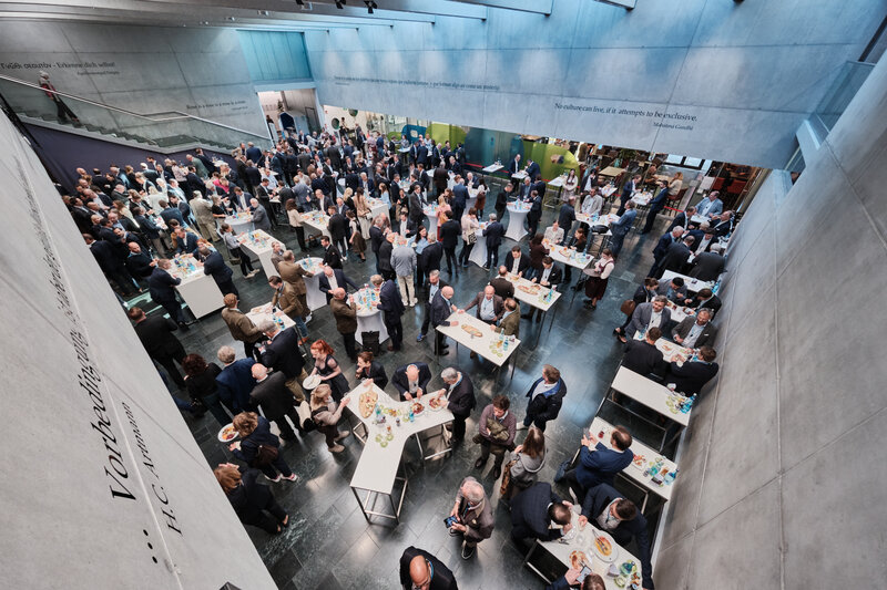 Blick von der Galerie in die lichtdurchflutete Aula der HOK: Die Teilnehmenden des 15. Internationalen Symposiums Restrukturierung tauschen sich beim Mittagsempfang an Stehtischen aus.