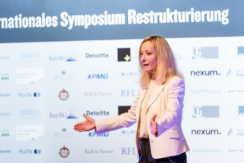 Dr. Romana Weber-Wilfert hält eine Präsentation im Veranstaltungsraum der HOK im Rahmen des 12. Internationalen Symposium Restrukturierung. Hinter ihr befindet sich eine Wand mit Sponsoring-Logos.